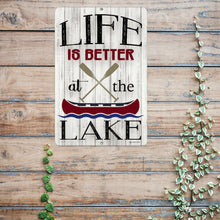 Load image into Gallery viewer, Detailed close-up of the Life Is Better at the Lake Metal Sign, showcasing bold typography, crossed paddles, and a vintage-style canoe illustration on a weathered wood background.
