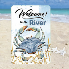Load image into Gallery viewer, Coastal-inspired Welcome to the River Metal Sign displayed on a sandy beach with seashells, adding rustic charm to riverfront homes, seafood restaurants, and beachside cottages.
