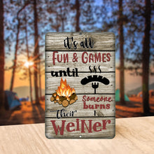 Load image into Gallery viewer, It&#39;s All Fun and Games Until Someone Burns Their Weiner Metal Sign styled outdoors at a scenic campsite, creating a fun and lighthearted camping vibe.
