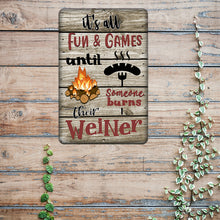 Load image into Gallery viewer, It&#39;s All Fun and Games Until Someone Burns Their Weiner Metal Sign displayed on a rustic wooden surface, adding a humorous and cozy touch to camping spaces.
