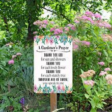 Load image into Gallery viewer, The Dyenamic Art Gardener’s Prayer sign mounted in a lush outdoor garden among pink flowering plants, illustrating its weather-resistant quality and perfect placement for gardeners and nature lovers.
