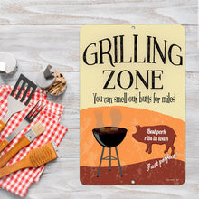Load image into Gallery viewer, Grilling Zone Metal Sign – Displayed on a wooden table with BBQ utensils and a checkered napkin, creating a fun ambiance.
