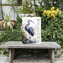 Load image into Gallery viewer, The Heron Welcome Metal Sign set in a lush outdoor garden, complementing greenery and floral accents. A serene and nature-inspired decorative piece for any space.
