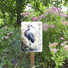 Load image into Gallery viewer, Heron Welcome Metal Sign mounted in a vibrant garden with blooming flowers, creating a peaceful and inviting entrance for outdoor spaces, patios, or pathways.
