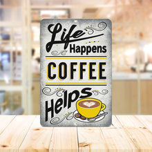 Load image into Gallery viewer, Life Happens Coffee Helps Metal Sign placed in a cozy coffee shop setting with wooden textures and warm lighting, adding a vintage coffeehouse vibe.

