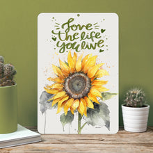Load image into Gallery viewer, Dyenamic Art Love The Life You Live Sunflower Metal Sign displayed on a shelf with potted plants, adding warmth to any space.
