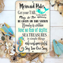 Load image into Gallery viewer, The Mermaid Rules Metal Sign displayed on a sandy beach with seashells and starfish, bringing a relaxing coastal vibe to any space. A decorative piece that adds character to beach houses and nautical-themed rooms.
