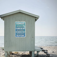 Load image into Gallery viewer, The Oceanology Metal Sign mounted on a rustic beach shack, blending seamlessly with the coastal scenery. A motivational and stylish wall sign that embodies the tranquility of ocean life, perfect for seaside homes and nautical lovers.
