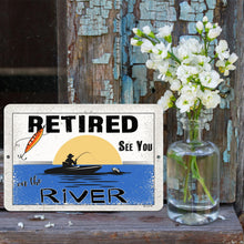 Load image into Gallery viewer, Dyenamic Art - Retired on the River Metal Sign – A Charming Fishing-Themed Retirement Gift Displayed Against a Rustic Wooden Backdrop with White Flowers. A Thoughtful and Sentimental Gift for Anglers.
