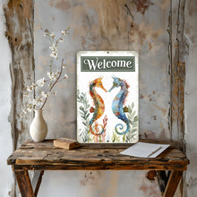 Load image into Gallery viewer, Seahorse Welcome Metal Sign displayed in a coastal-themed setting with tropical greenery, seashells, and a sandy background, creating a serene and inviting beach aesthetic.
