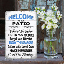 Load image into Gallery viewer, The  Welcome to Our Patio metal sign displayed against a rustic wooden wall with a vase of white flowers, perfect for porch or deck decor.
