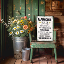 Load image into Gallery viewer, A rustic white wooden sign with black text reading 'FARMHOUSE RULES' is propped against a distressed green wooden chair on a porch. The chair sits beside a galvanized metal bucket filled with daisies, sunflowers, and other wildflowers, with a small metal watering can nearby.
