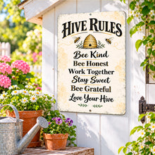Load image into Gallery viewer, Rustic Hive Rules bee sign displayed against a light garden wall surrounded by colorful flowers, terracotta pots, and lush greenery.
