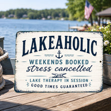 Load image into Gallery viewer, Lakeaholic metal sign displayed on a lakeside dock table overlooking the water with lake house decor and scenic waterfront background
