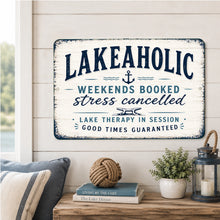 Load image into Gallery viewer, Lakeaholic metal sign displayed in a cozy lake house living room with rustic decor, lake-themed books, lantern, and waterfront view
