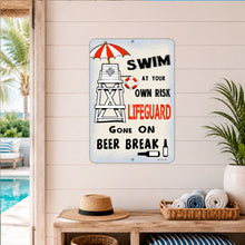 Load image into Gallery viewer, A rectangular metal wall sign with a beach-themed warning message reads 'SWIM AT YOUR OWN RISK LIFEGUARD GONE ON BEER BREAK' with an illustrated lifeguard tower, red-and-white umbrella, and small beer glass. The sign is displayed on a light beige shiplap wall above a wooden console table with a straw hat, rolled towels, and a potted plant, with a pool and palm trees visible in the background.
