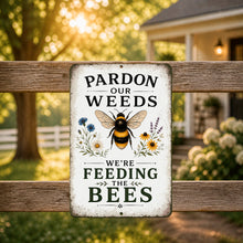 Load image into Gallery viewer, Pardon the Weeds Feeding Bees metal garden sign displayed on rustic fence with warm cottage garden background and pollinator-friendly decor theme
