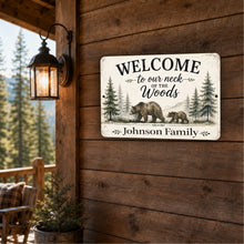 Load image into Gallery viewer, A rustic rectangular metal sign mounted on a wooden cabin wall, featuring the text “WELCOME to our neck of the Woods” and “Johnson Family” with illustrated bears, trees, and mountains. A lit wall lantern and outdoor porch setting are visible beside it.
