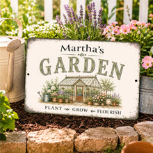 Load image into Gallery viewer, Personalized garden sign displayed outdoors distressed cream background reading 'Martha's GARDEN' and 'PLANT GROW FLOURISH,' featuring an illustrated greenhouse and plants. The sign is photographed outdoors in a garden setting with flowers, mulch, stones, and metal garden tools around it.
