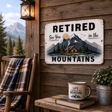 Load image into Gallery viewer, Retired sign with mountain scene on wooden cabin wall, mug, book, and plant on table.
