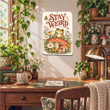 Load image into Gallery viewer, Stay Weird Frog Metal Wall Sign displayed in a cozy cottagecore room with rustic wooden furniture, botanical accents, greenery, and warm natural lighting.
