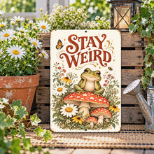 Load image into Gallery viewer, Stay Weird Frog Metal Sign leaning against a rustic wooden crate surrounded by daisies, garden plants, and charming cottagecore decorations.
