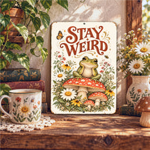Load image into Gallery viewer, Whimsical Stay Weird Frog Metal Sign displayed in a cozy reading nook with vintage books, daisies, rustic wooden accents, and cottagecore decor.
