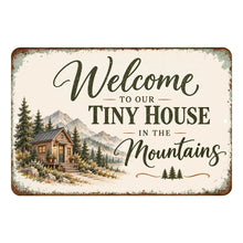 Load image into Gallery viewer, Horizontal tiny house welcome metal sign with a rustic mountain cottage, evergreen forest, green lettering, and weathered antique-style edges on a white background.
