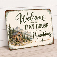 Load image into Gallery viewer, Rustic aluminum welcome sign featuring a small timber cabin surrounded by pine trees and mountains, photographed at an angle against a light background.
